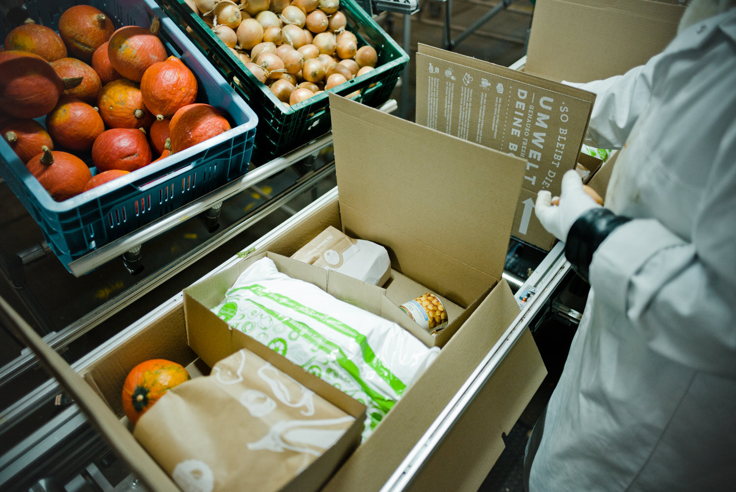 Mitarbeiter in Handschuhen packt Zutaten in eine HelloFresh-Box, daneben Kisten mit Kürbissen und Zwiebeln